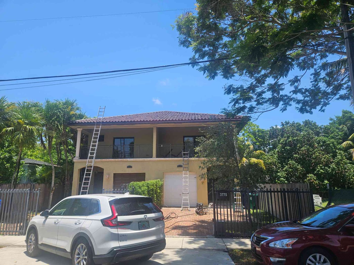 Tan two-story stucco home with a barrel-tile roof and balcony mid-repaint, ladders set against the front facade