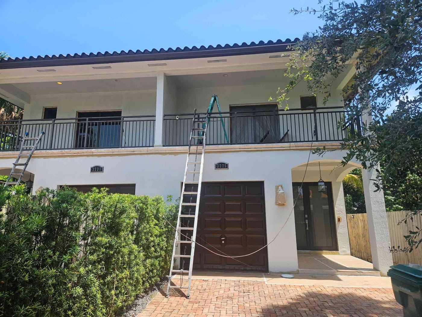 White two-story home with a dark brown garage door and balcony railing during an exterior repaint, ladder in the driveway