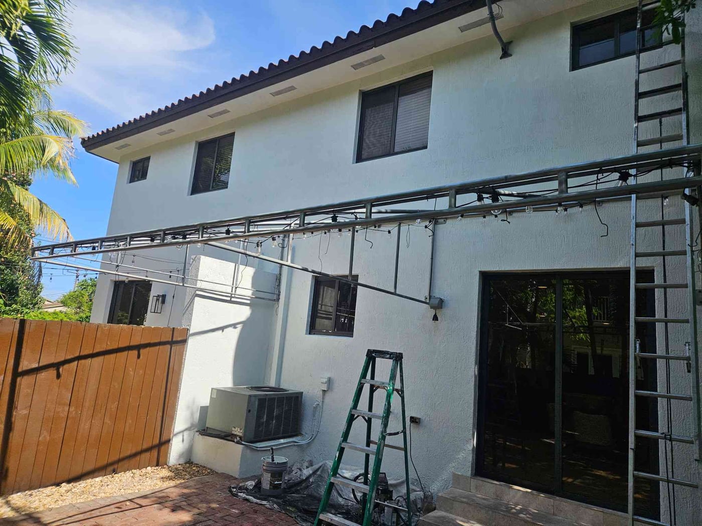Rear of a white two-story home being repainted, with a steel pergola frame and air-conditioning unit