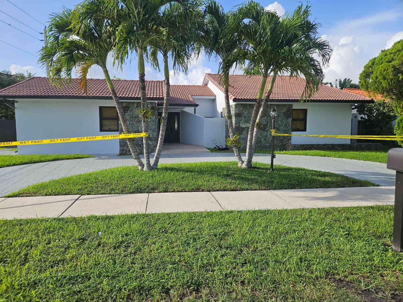 Freshly painted single-story home with stone accents and palm trees, wet-paint tape across the front yard