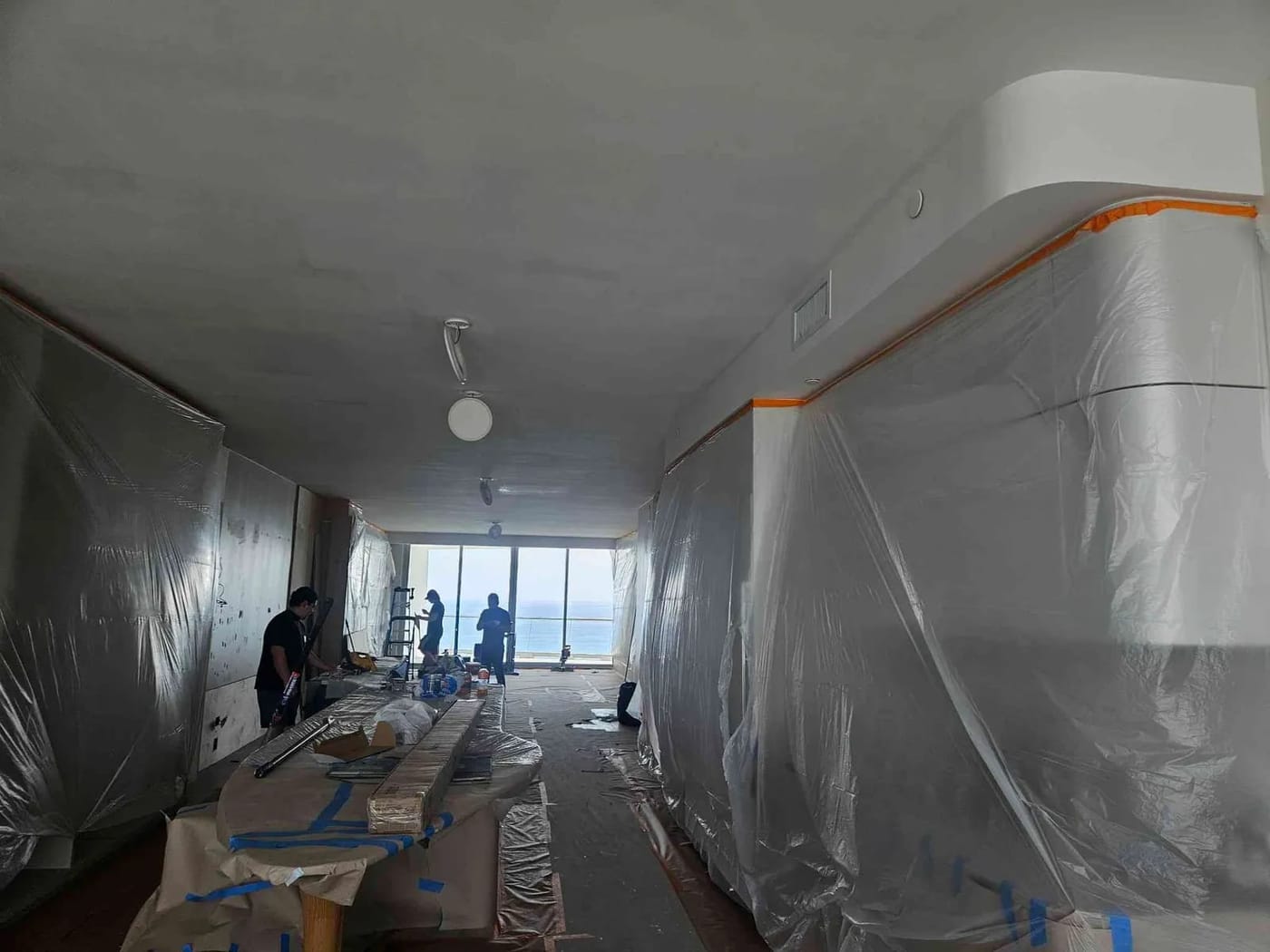 High-rise oceanfront condo interior prepped with plastic sheeting for painting, with painters working by the ocean-view windows