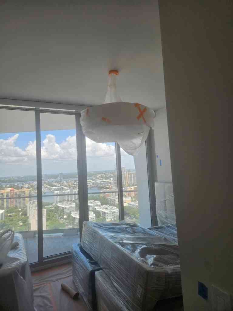 Condo bedroom with floor-to-ceiling windows over the coastline, light fixture bagged and furniture wrapped for painting