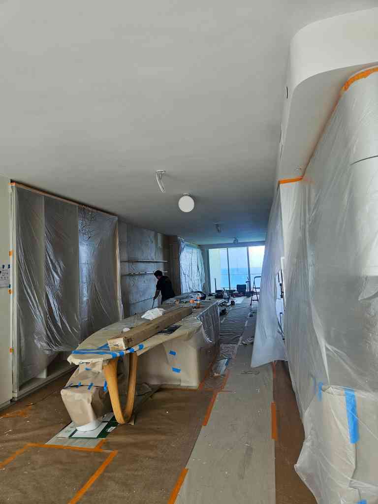 Oceanfront condo interior masked with plastic sheeting during a repaint, painter working near the window wall