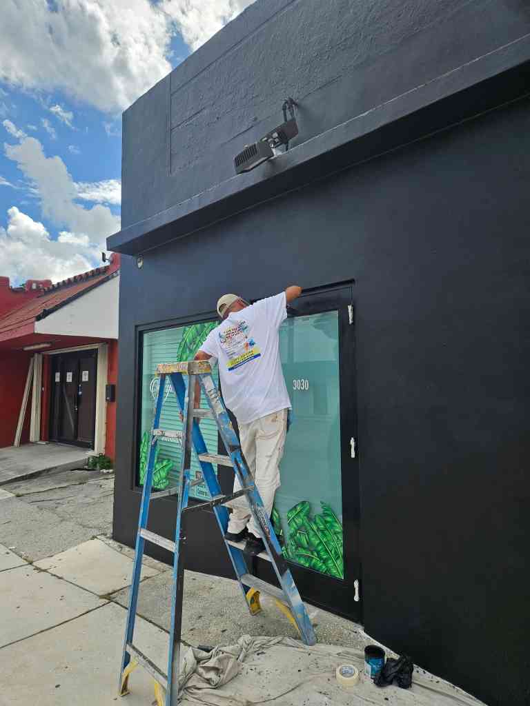 Painter on a ladder coating the black exterior of a commercial storefront with a leaf-mural glass door