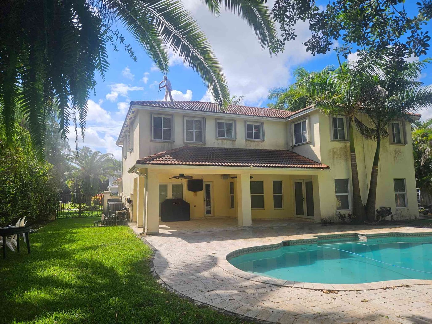 Two-story tan home with a barrel-tile roof beside a backyard pool surrounded by palms