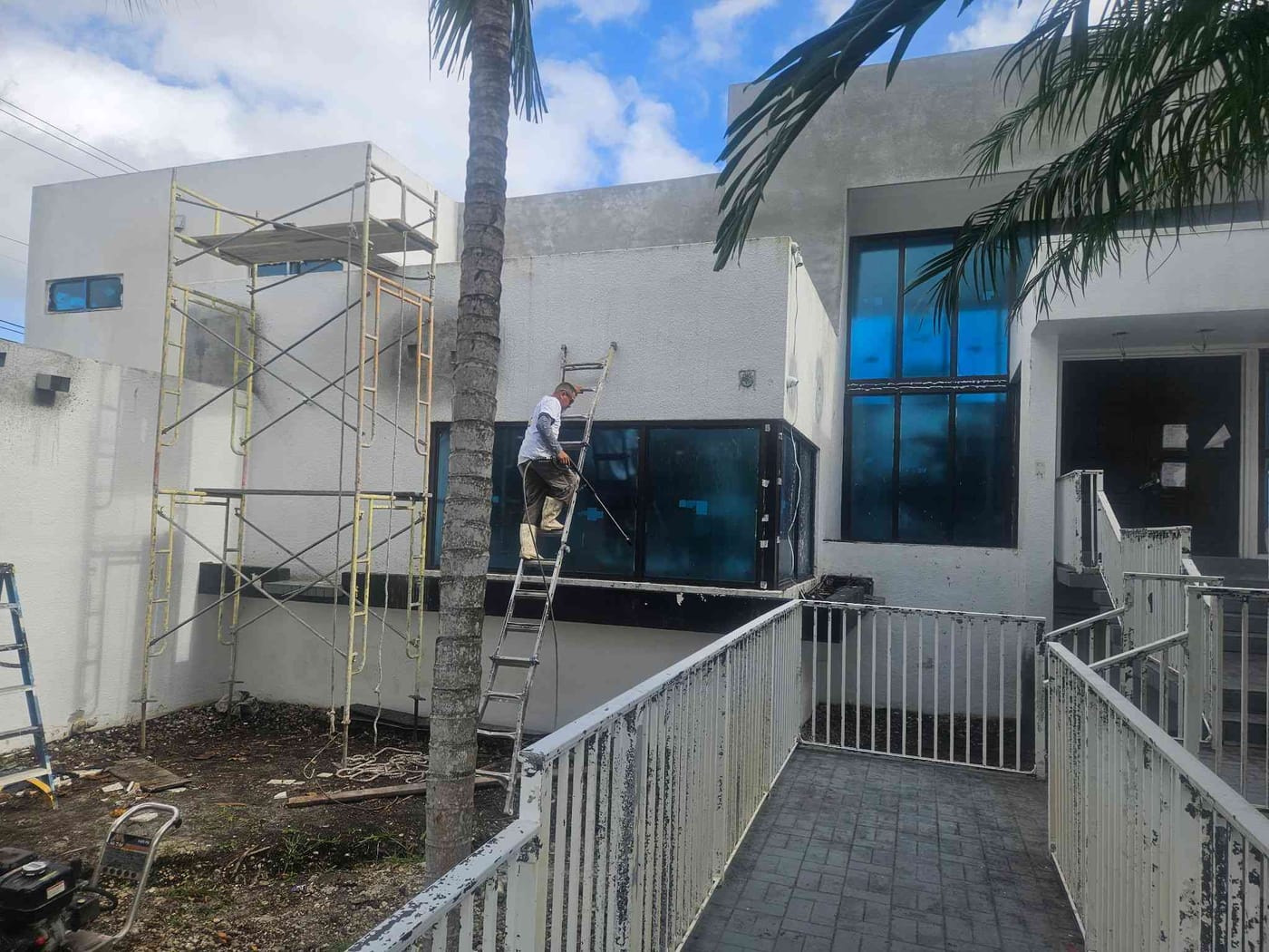 Painter on a ladder beside scaffolding working on a two-story building's exterior near a white railing