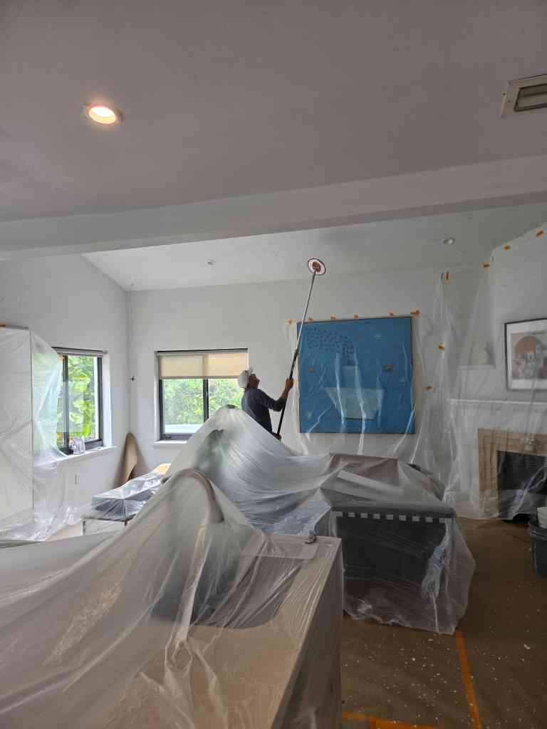Painter using a roller extension to repaint a living-room ceiling, with furniture protected under plastic sheeting